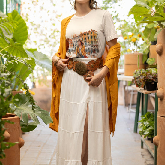 A person stands in a greenhouse wearing a tan hat, Bella Vita Voyager Tee with a city print, long white skirt with a slit, and tan shoes. They drape an organic cotton yellow-orange shawl and an ornate belt. Green plants surround them as natural light filters in from above.