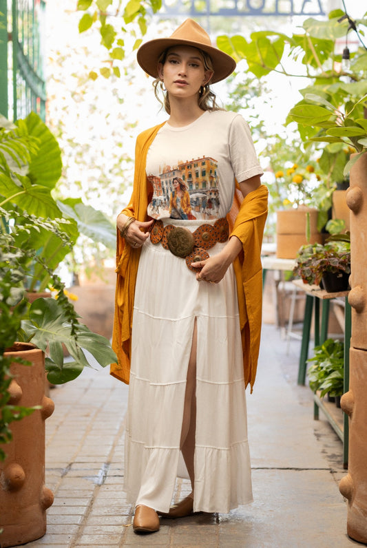 A person stands in a greenhouse wearing a tan hat, Bella Vita Voyager Tee with a city print, long white skirt with a slit, and tan shoes. They drape an organic cotton yellow-orange shawl and an ornate belt. Green plants surround them as natural light filters in from above.