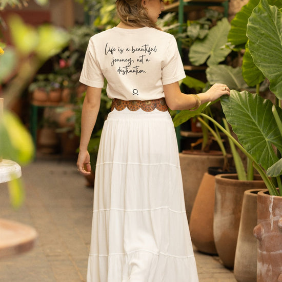 A woman walks down a plant-filled corridor, barefoot in an eco-conscious Bella Vita Voyager Tee, featuring the phrase "Life is a beautiful journey, not a destination." Her tan hat and flowing white skirt complete the peaceful scene as she moves among large potted plants and clay pots. It's truly a Bella Vita Voyageur experience.