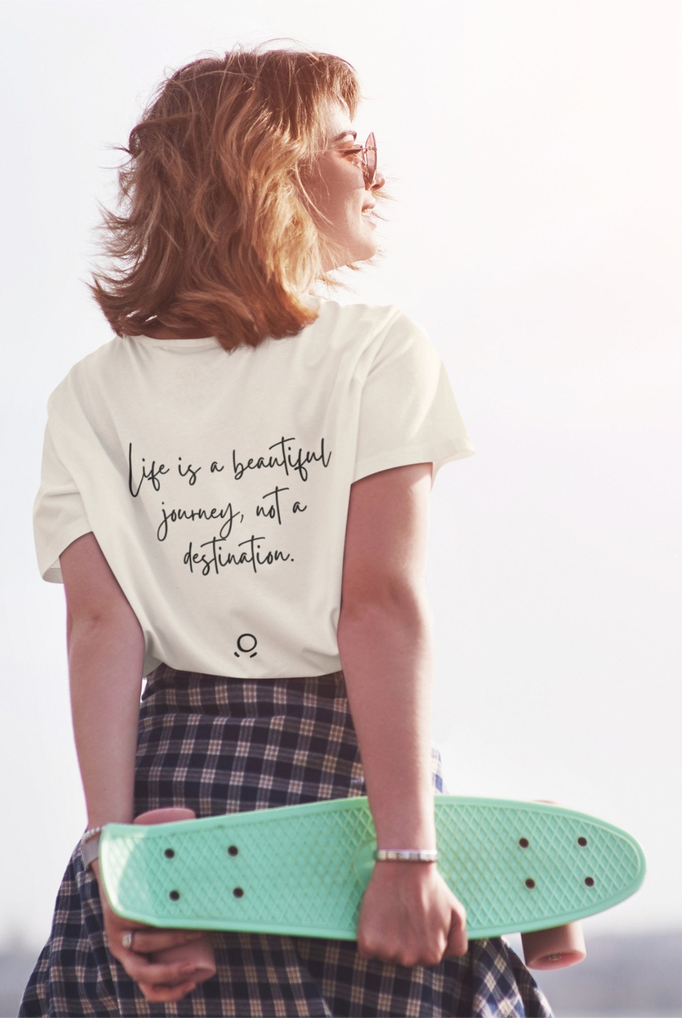 A person with short hair holds a turquoise skateboard behind their back. They wear a Bella Vita Voyager Tee in white, which features the quote, "Life is a beautiful journey, not a destination," along with a plaid skirt. The person smiles as they gaze toward the sunlight, creating an uplifting and vibrant scene.