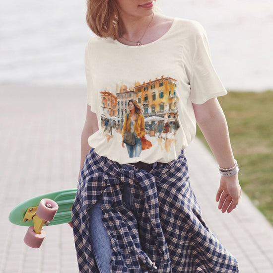 A woman strolls outdoors with a skateboard, wearing sunglasses, a Bella Vita Voyager Tee, jeans, and a plaid shirt tied at her waist. Her shoulder-length hair frames a soft smile. In the background, blurred water and a pathway hint at her adventurous spirit.