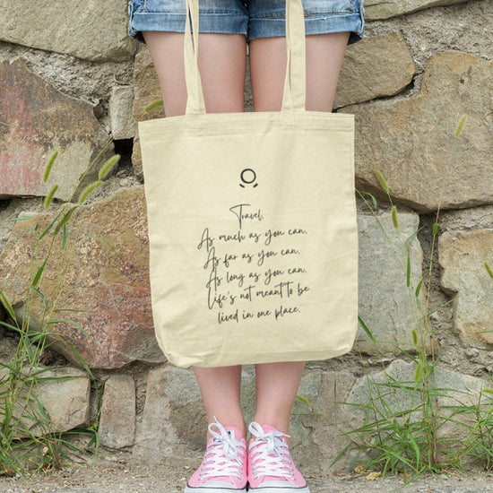 A person leans against a stone wall, wearing denim shorts and pink sneakers, holding the eco-friendly Horizon Seeker Tote Bag. This beige bag features the empowering message in black text: "Travel as much as you can, as far as you can, as long as you can. Life’s not meant to be lived in one place.