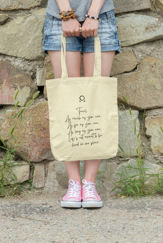A person leans against a stone wall, wearing denim shorts and pink sneakers, holding the eco-friendly Horizon Seeker Tote Bag. This beige bag features the empowering message in black text: "Travel as much as you can, as far as you can, as long as you can. Life’s not meant to be lived in one place.