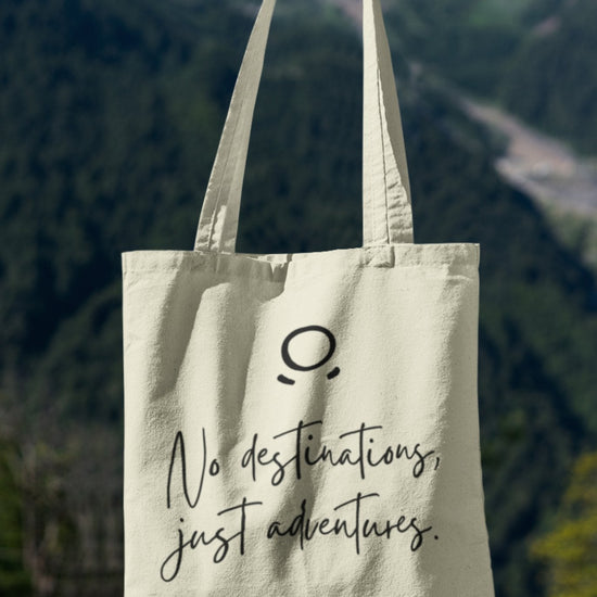 A hand holds the Adventure Awaits Tote Bag against a mountain backdrop. This eco-friendly tote bag showcases an elegant minimalist design with a small circle above a curved line, resembling a face, and is inscribed with the cursive message "No destinations, just adventures." Adventure Awaits!