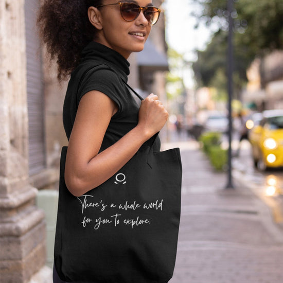 A woman with sunglasses and curly hair stands on a city sidewalk, smiling brightly. Embracing her adventurous spirit, she dons a black sleeveless top and blue pants while carrying an eco-friendly Cosmic Voyager Tote Bag emblazoned with the message, "There's a whole world for you to explore." Behind her, trees line the street where a yellow car is parked.