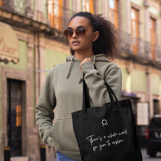A woman with an adventurous soul, wearing sunglasses and a green hoodie, holds a Cosmic Voyager Tote Bag that reads "There's a whole world for you to explore." She stands on a city street with historic buildings in the background, featuring ornate balconies and large windows.
