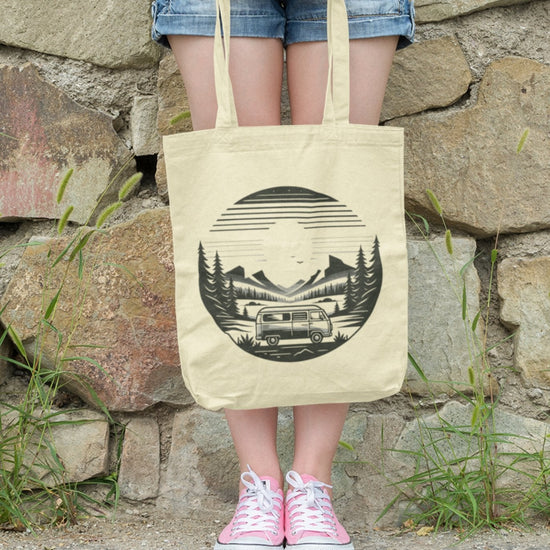 A person stands against a stone wall with grass protruding from cracks, holding the Horizon Seeker Tote Bag—an eco-friendly accessory featuring a black design of a van, trees, and mountain under the sun. They wear a gray shirt, denim shorts, and pink sneakers.
