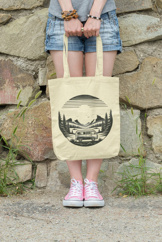 A person stands against a stone wall with grass protruding from cracks, holding the Horizon Seeker Tote Bag—an eco-friendly accessory featuring a black design of a van, trees, and mountain under the sun. They wear a gray shirt, denim shorts, and pink sneakers.