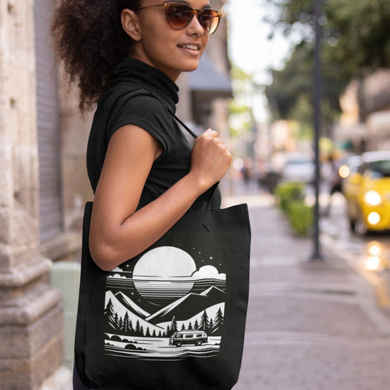 A woman wearing sunglasses and a black turtleneck smiles as she strolls down a city street, carrying the Cosmic Voyager Tote Bag. This eco-friendly tote showcases a white graphic of a mountainous landscape with trees, a river, and a large sun or moon. In the background, there is a blurred yellow car and some greenery.