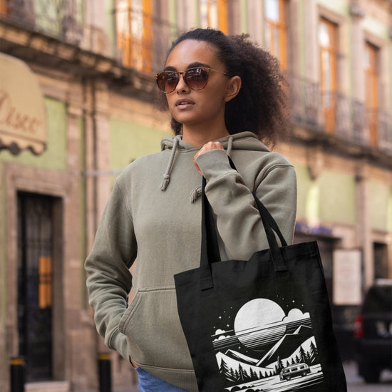 A person wearing sunglasses and a green hoodie strolls down a city street, embodying an adventurous soul. They carry the Cosmic Voyager Tote Bag, adorned with mountains, trees, and a moon. Behind them, a building with green walls and ornate balconies completes the scene.