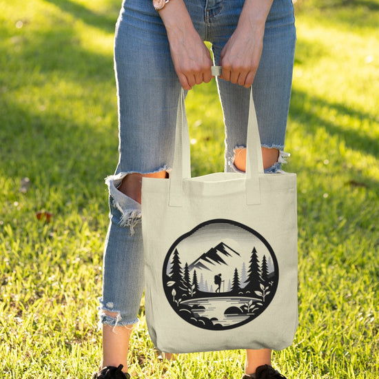 A person stands on grass holding the Adventure Awaits Tote Bag, featuring a mountain and forest silhouette design. They wear a light pink top, ripped blue jeans, black sneakers, and a watch. The sun casts long shadows, suggesting late afternoon—perfect for sustainable exploration.