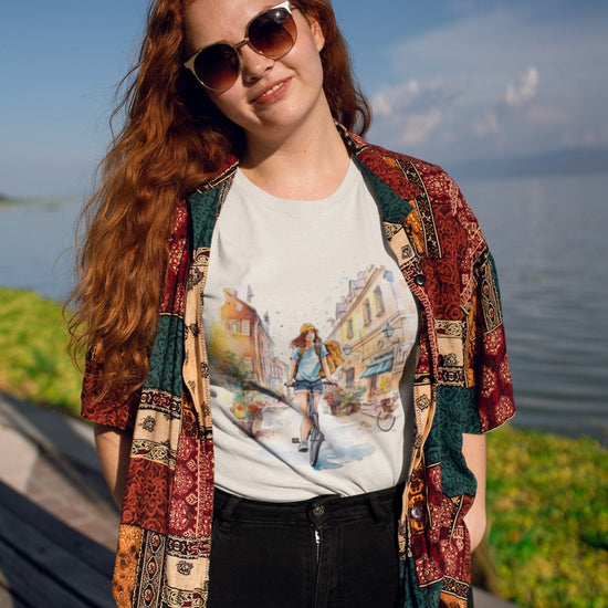 A woman with long red hair is by the lakeside under a clear sky, wearing sunglasses and a colorful patterned shirt layered over an eco-friendly "Pedal through Life" T-shirt that features a street scene design. Soft sunlight casts gentle shadows, highlighting the lush greenery and tranquil water that mirror her commitment to sustainable living.