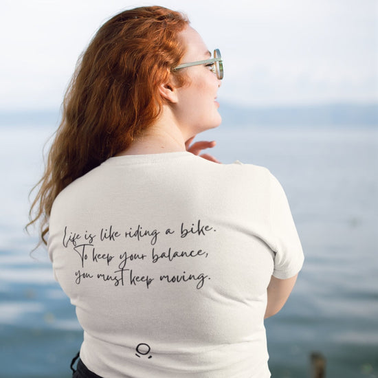 A woman with long red hair stands by a tranquil lake, facing away from the camera. She wears sunglasses and a Pedal through Life Tee, crafted from organic cotton for an eco-friendly touch. The shirt displays the text, "Life is like riding a bike. To keep your balance, you must keep moving." Above her is a clear sky, and in front of her stretches a wooden dock into the water.