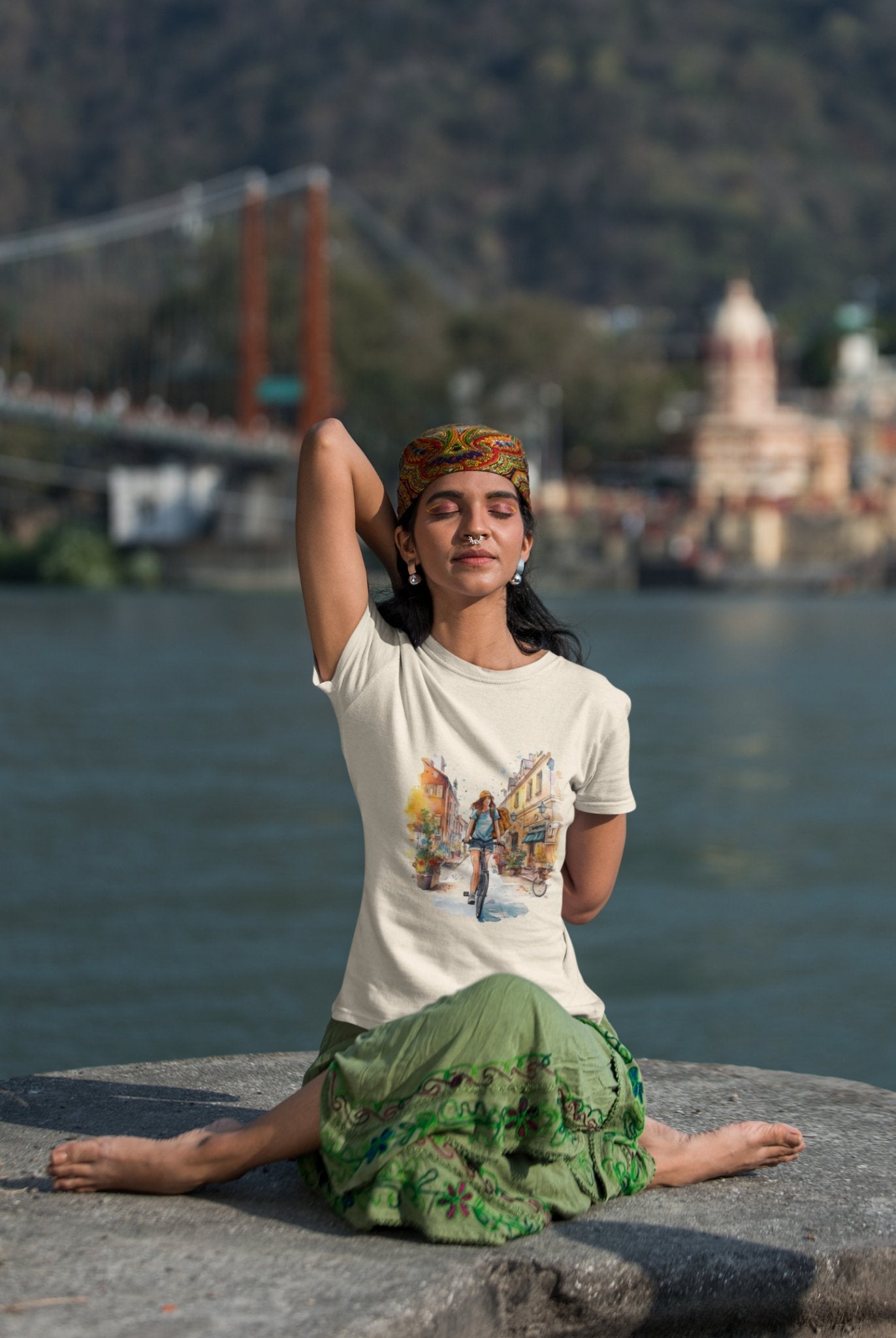 A person sits cross-legged on a stone ledge by a river, performing a yoga pose with one arm raised. They wear a colorful headband, the eco-friendly "Pedal through Life Tees," and a green skirt. In the background, there's a suspension bridge and blurred buildings amid a hilly landscape.