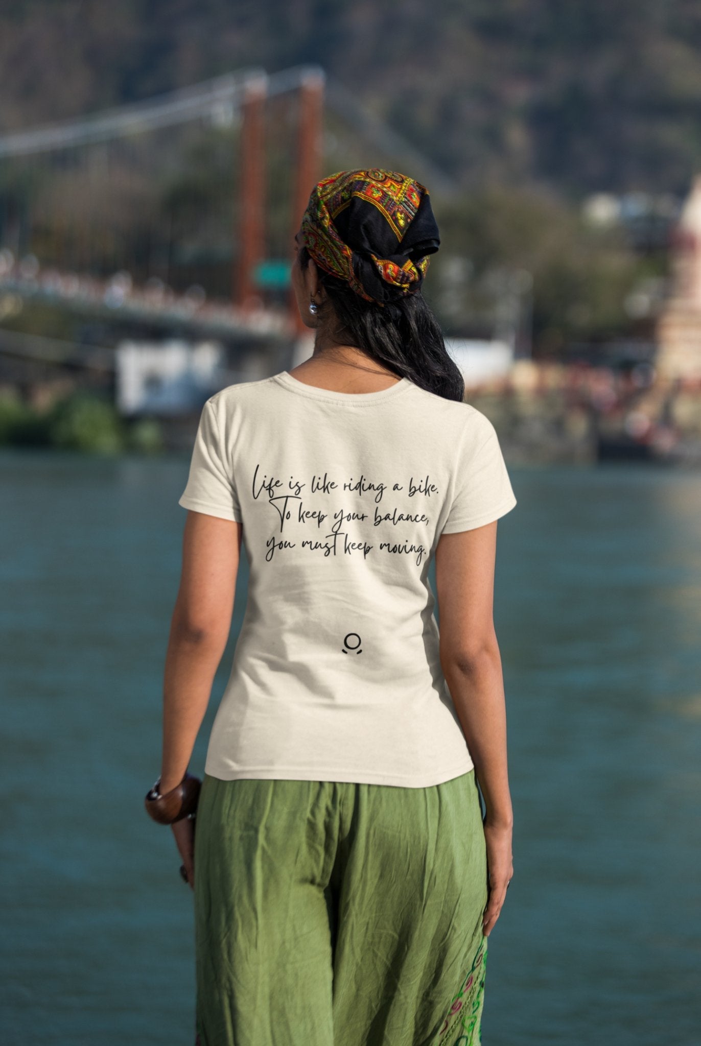 A person stands by a river wearing the eco-friendly "Pedal through Life" T-shirt featuring the quote, "Life is like riding a bike. To keep your balance, you must keep moving." The individual has long hair partially covered by a patterned scarf and wears green pants. A bridge and blurred trees are visible in the background.