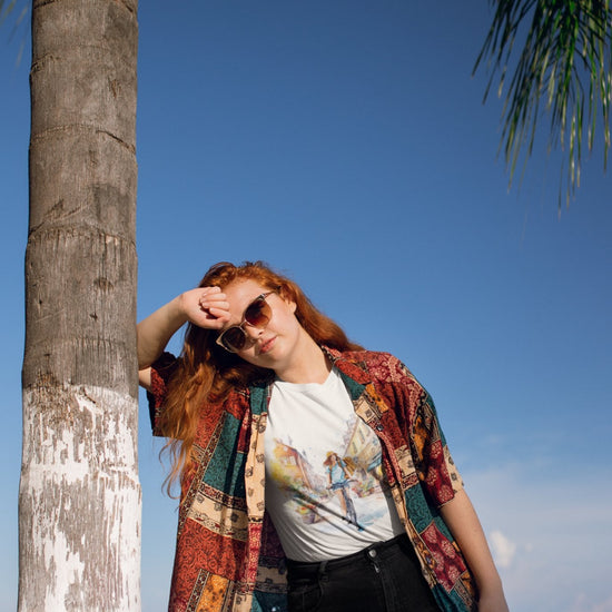 A person with long red hair leans casually against a palm tree, exuding cool vibes in their sunglasses and colorful patchwork shirt. They sport the "Pedal through Life" tee made from organic cotton. Against the backdrop of a clear blue sky and water, palm leaves sway gently at the top right corner.