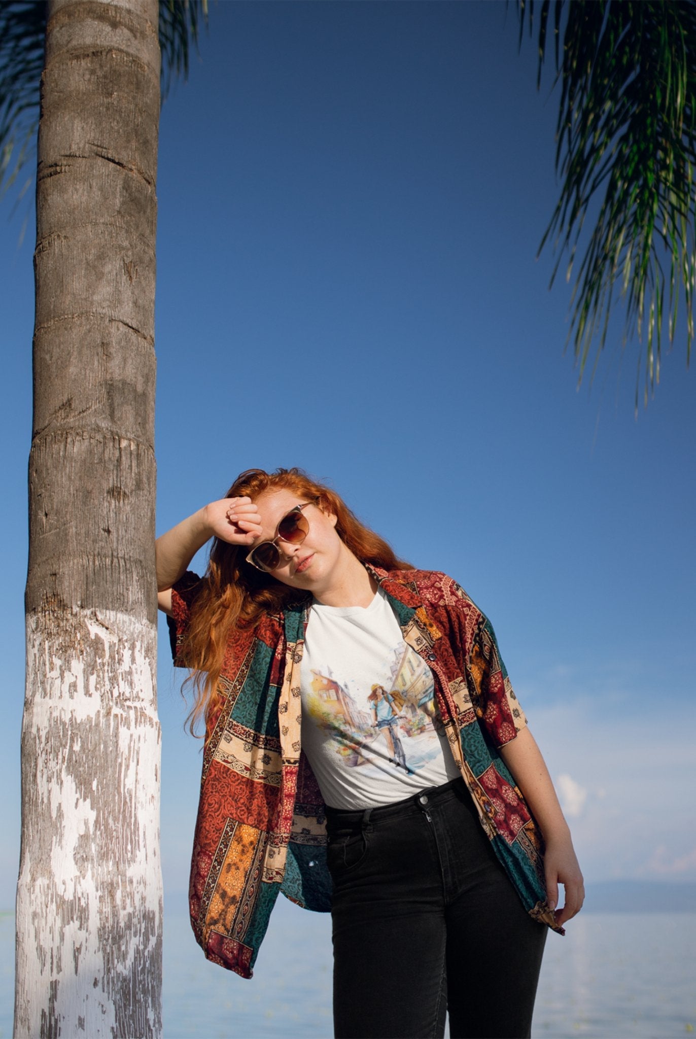 A person with long red hair leans casually against a palm tree, exuding cool vibes in their sunglasses and colorful patchwork shirt. They sport the "Pedal through Life" tee made from organic cotton. Against the backdrop of a clear blue sky and water, palm leaves sway gently at the top right corner.