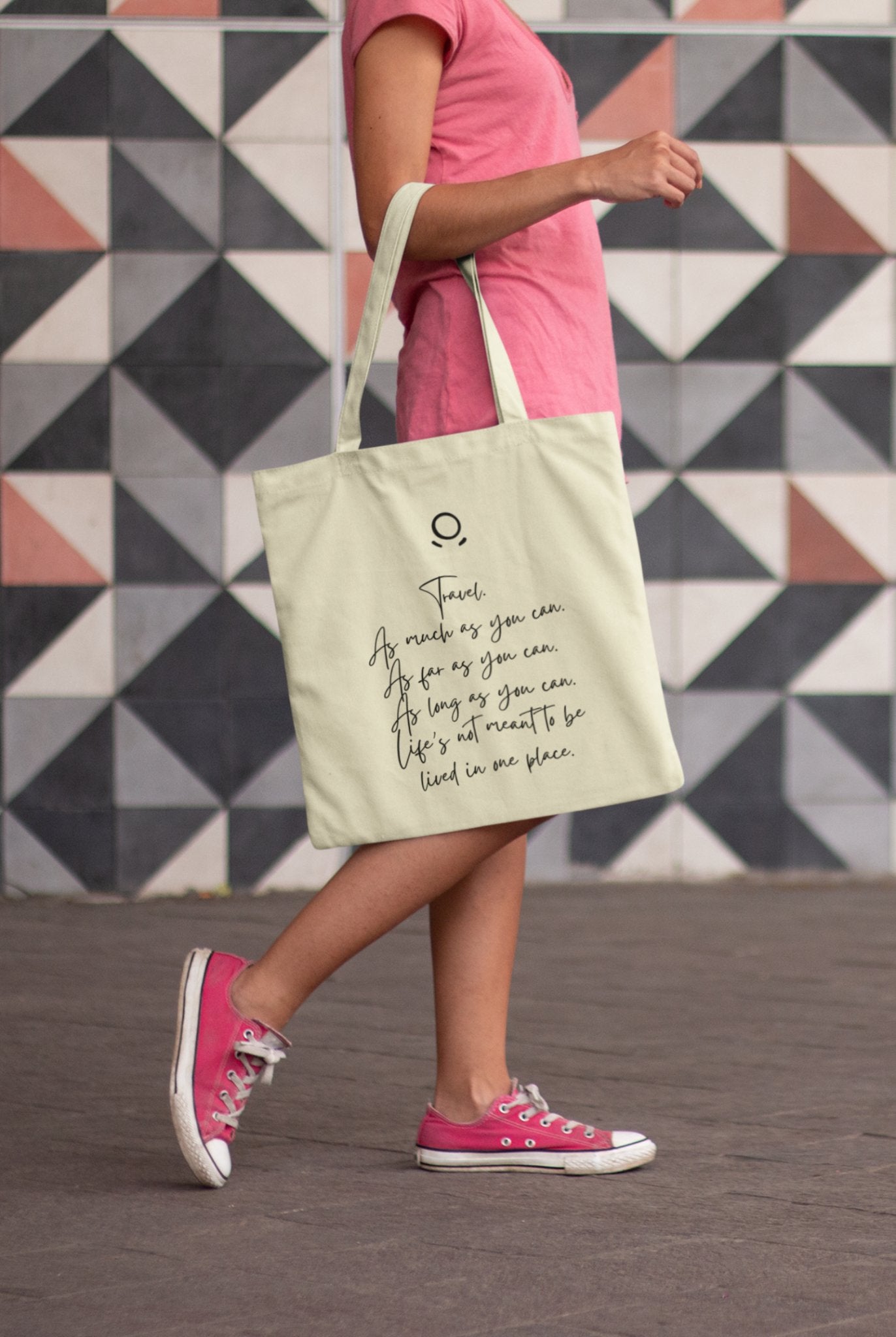 A person in a pink shirt and sneakers walks with the Horizon Seeker Tote Bag, an eco-friendly, cream-colored bag displaying a quote in black cursive. The background features gray and white geometric tiles.