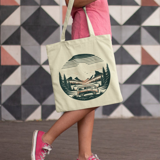 A person wearing a pink t-shirt, pink sneakers, and denim shorts carries the eco-friendly Horizon Seeker Tote Bag featuring a vintage trailer in a forest scene. The backdrop displays black, white, and gray geometric patterned tiles.