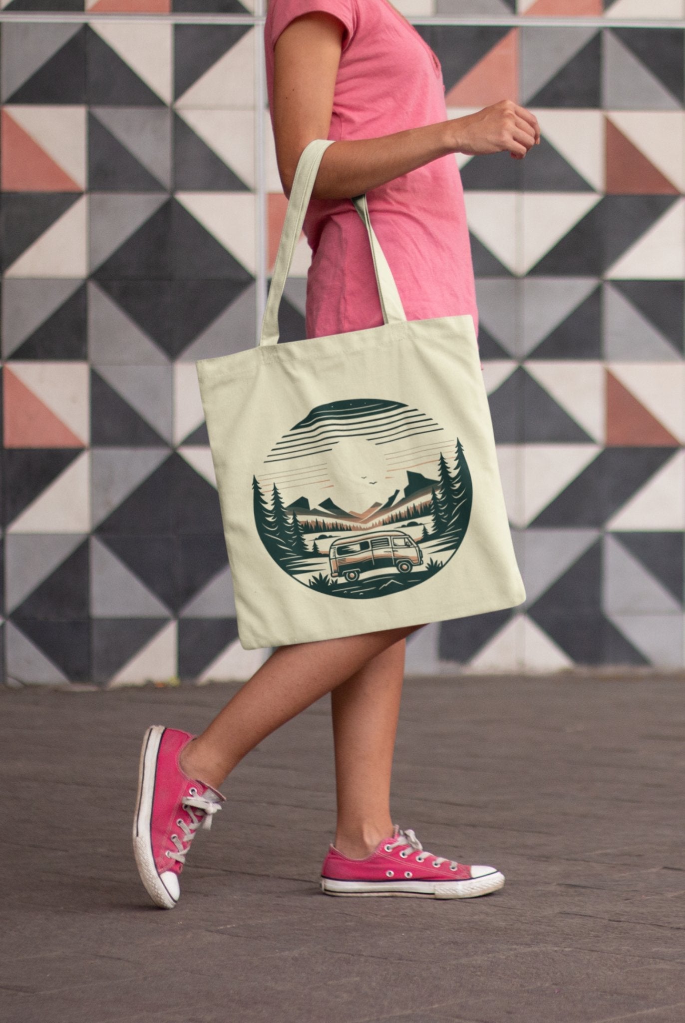 A person wearing a pink t-shirt, pink sneakers, and denim shorts carries the eco-friendly Horizon Seeker Tote Bag featuring a vintage trailer in a forest scene. The backdrop displays black, white, and gray geometric patterned tiles.