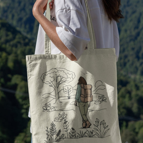 An adventurous woman stands outside, clutching her eco-friendly The Adventurous Woman Tote Bag, decorated with a design of a hiker exploring a natural setting surrounded by trees and shrubs. She is wearing a white shirt and jeans against the backdrop of forested hills.
