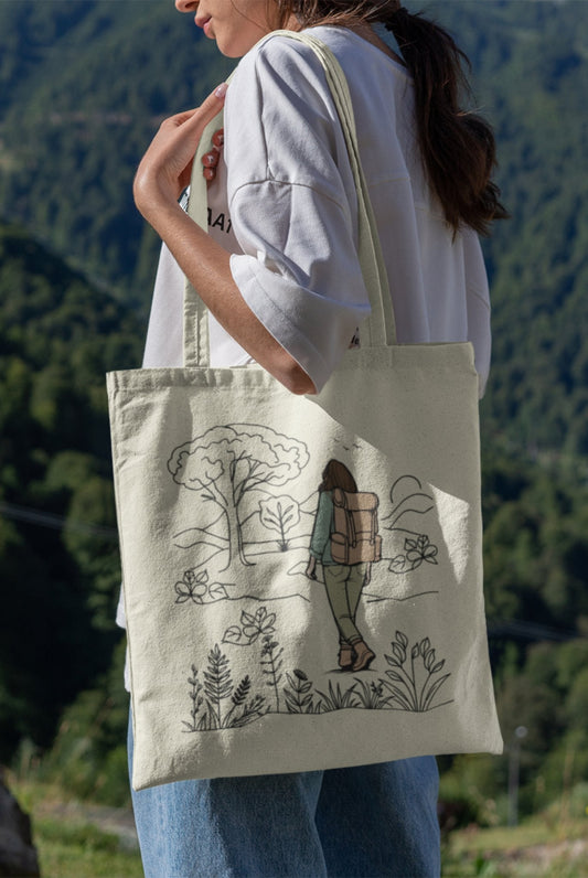 An adventurous woman stands outside, clutching her eco-friendly The Adventurous Woman Tote Bag, decorated with a design of a hiker exploring a natural setting surrounded by trees and shrubs. She is wearing a white shirt and jeans against the backdrop of forested hills.
