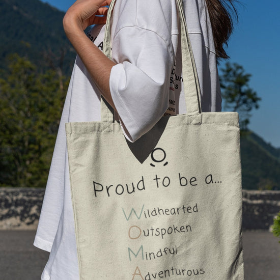 A person holds "The Adventurous Woman Tote Bag," an eco-friendly, cream-colored accessory adorned with the colorful and empowering message: "Proud to be an Adventurous Woman." Each letter symbolizes being Wildhearted, Outspoken, Mindful, Adventurous, and Nurturing. Dressed in a white shirt and jeans, they are outdoors with nature softly fading into the background.