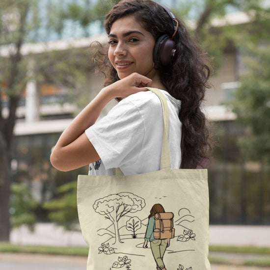 A woman with curly hair, donned in a white t-shirt, smiles as she wears headphones and holds "The Adventurous Woman Tote Bag." This eco-friendly beige tote, ideal for sustainable travel, showcases a design of a person with a backpack exploring nature. She stands on a city sidewalk with trees and blurred buildings in the background.