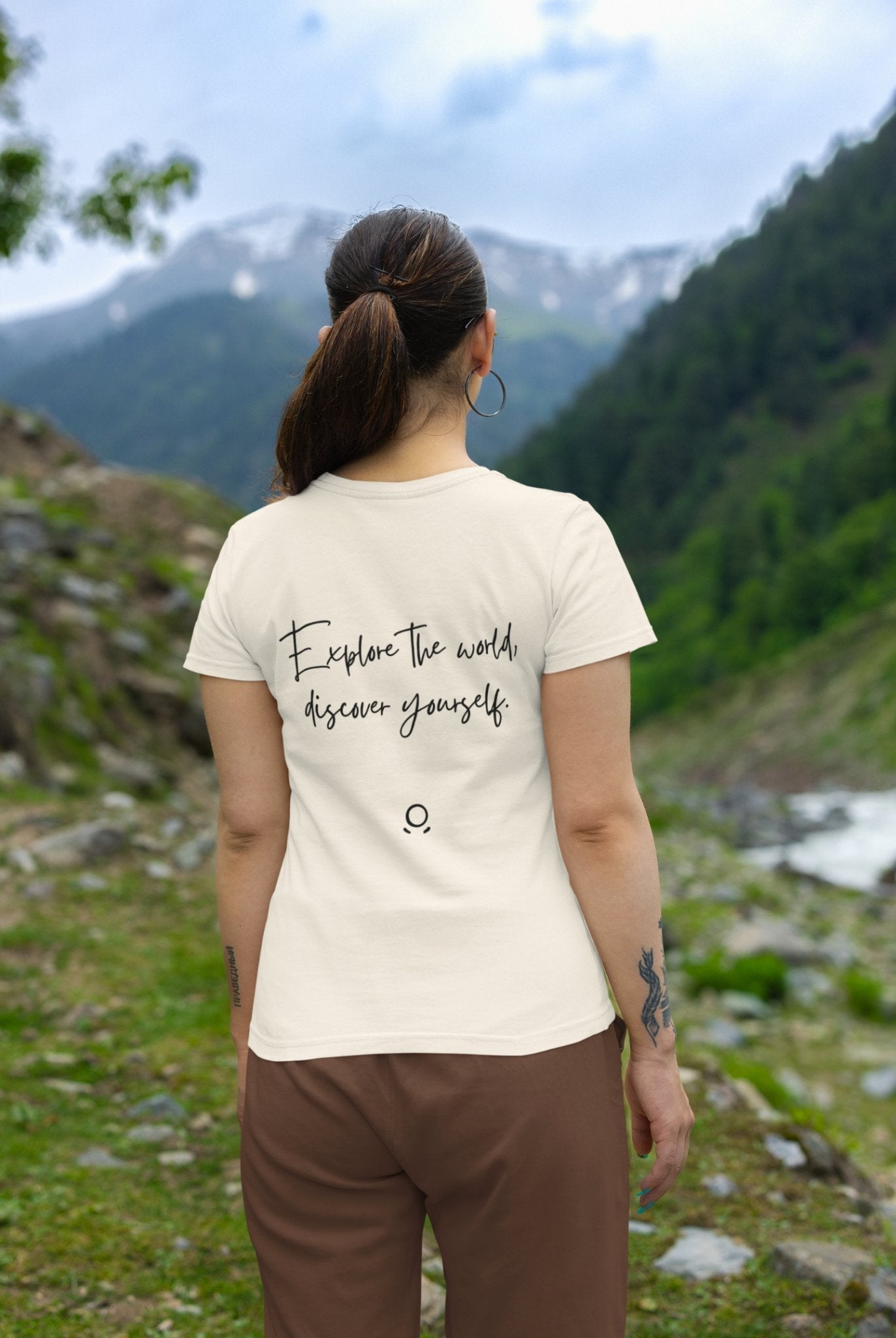 A woman with a ponytail stands outdoors, her eco-conscious Transit Trailblazer Tee adorned with the phrase "Explore the world, discover yourself" as she gazes upon lush green hills and mountains under a cloudy sky. Her spirit of wanderlust perfectly aligns with the breathtaking scenery.