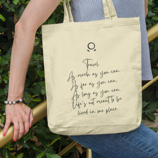 Clad in a sleeveless gray top and blue jeans, a person carries the eco-friendly Horizon Seeker Tote Bag over their shoulder. The beige tote with black script text—"Travel. As much as you can. As far as you can. As long as you can."—contrasts the lush green backdrop.