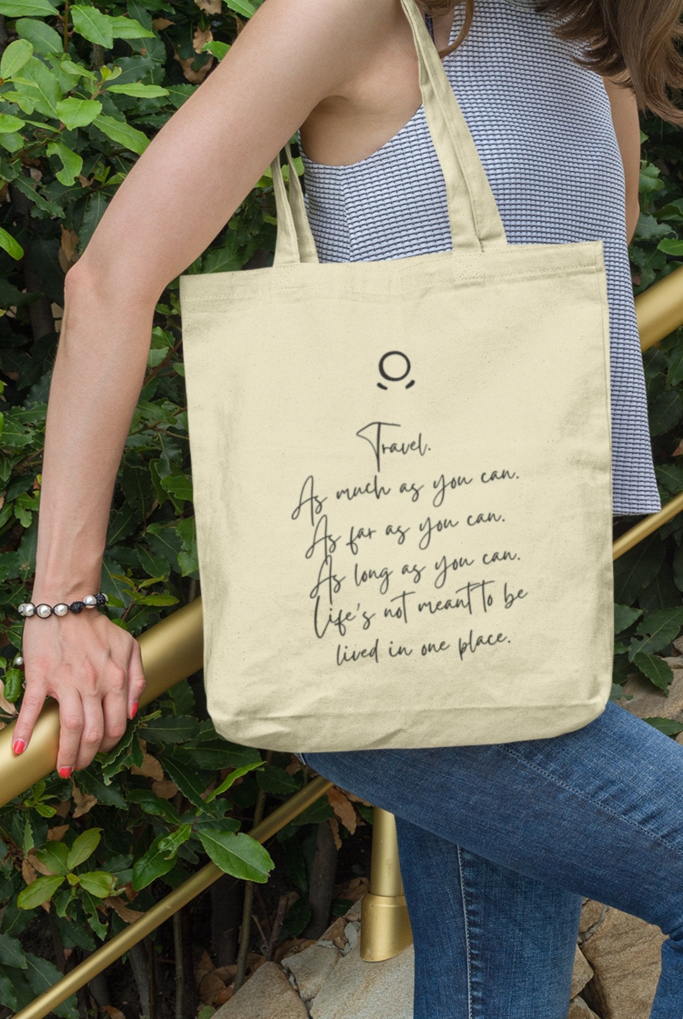 Clad in a sleeveless gray top and blue jeans, a person carries the eco-friendly Horizon Seeker Tote Bag over their shoulder. The beige tote with black script text—"Travel. As much as you can. As far as you can. As long as you can."—contrasts the lush green backdrop.