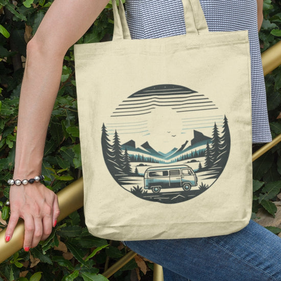 A person in a striped tank top and blue jeans holds the eco-friendly Horizon Seeker Tote Bag, featuring a camper van, forest, mountains, and setting sun design. Standing near greenery, they grasp a golden railing.