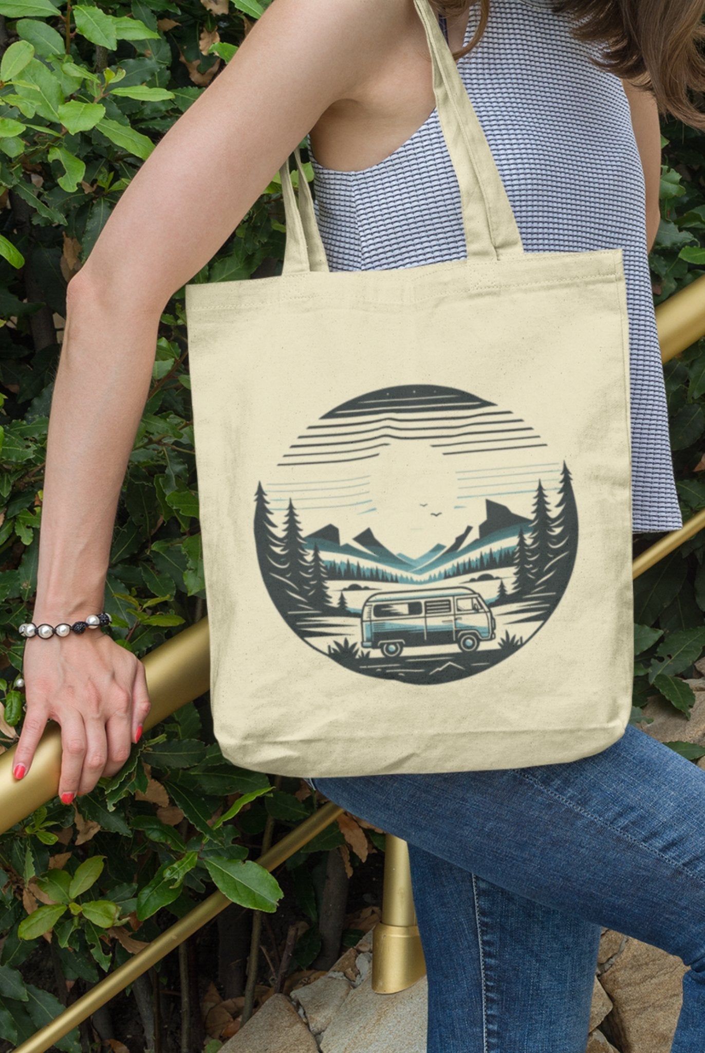 A person in a striped tank top and blue jeans holds the eco-friendly Horizon Seeker Tote Bag, featuring a camper van, forest, mountains, and setting sun design. Standing near greenery, they grasp a golden railing.