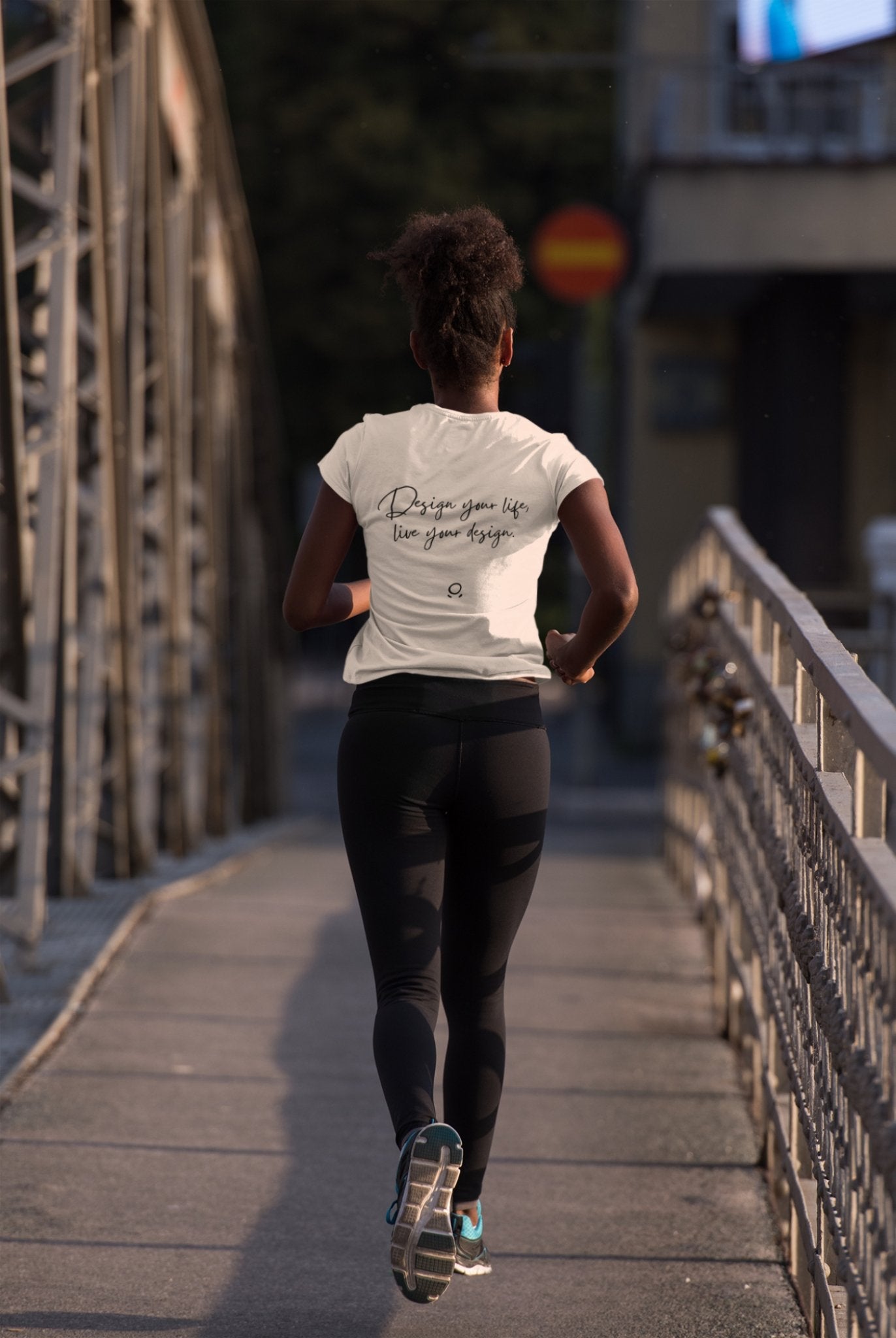 From behind, a person is seen jogging on a bridge, embodying their wanderlust in a white "Wanderlust Workspace" tee that says "Design your life like your design," paired with black leggings. The eco-conscious bridge features metal railings and a paved path, beautifully illuminated by the gentle light of early morning or late afternoon.