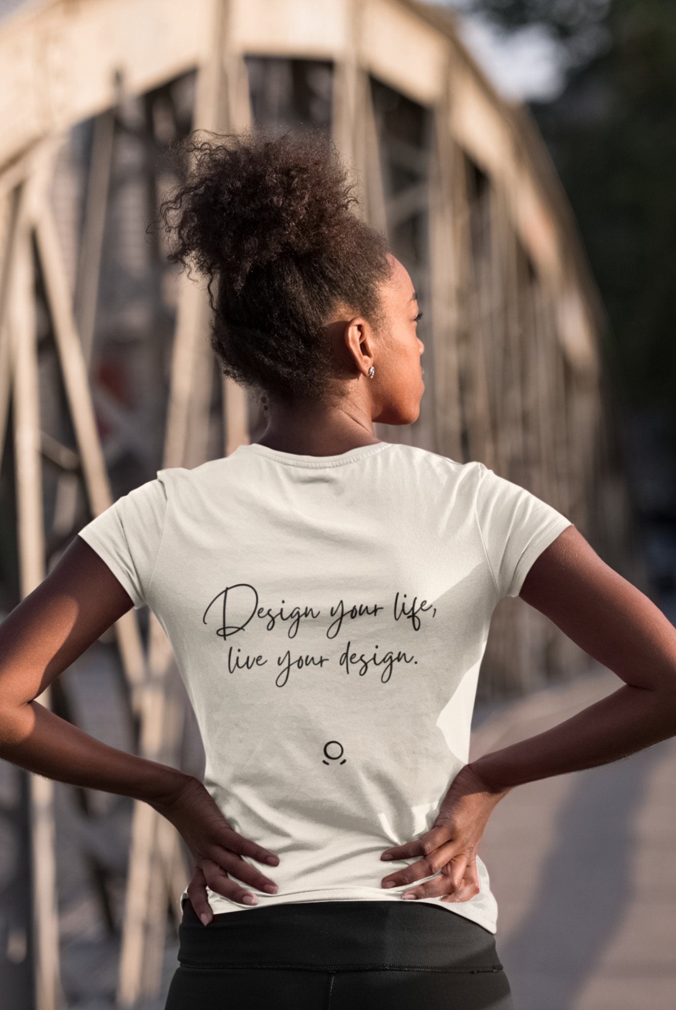 A woman with curly hair styled in a bun stands confidently on a metal bridge, wearing the Wanderlust Workspace Tee, which says "Design your life, live your design." Channeling the essence of digital nomads and an eco-friendly lifestyle, she gazes away from the camera with her hands on her hips.