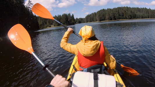 Two people kayaking on a serene lake, surrounded by lush forests. The person in the foreground wears an eco-friendly orange jacket and yellow bandana, highlighting sustainable outdoor adventure apparel.