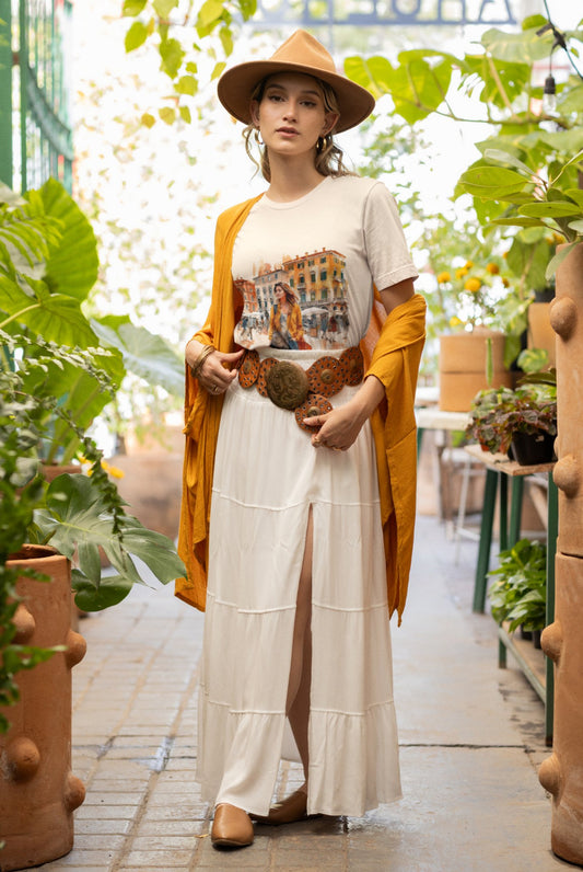 A person stands in a greenhouse wearing a tan hat, Bella Vita Voyager Tee with a city print, long white skirt with a slit, and tan shoes. They drape an organic cotton yellow-orange shawl and an ornate belt. Green plants surround them as natural light filters in from above.