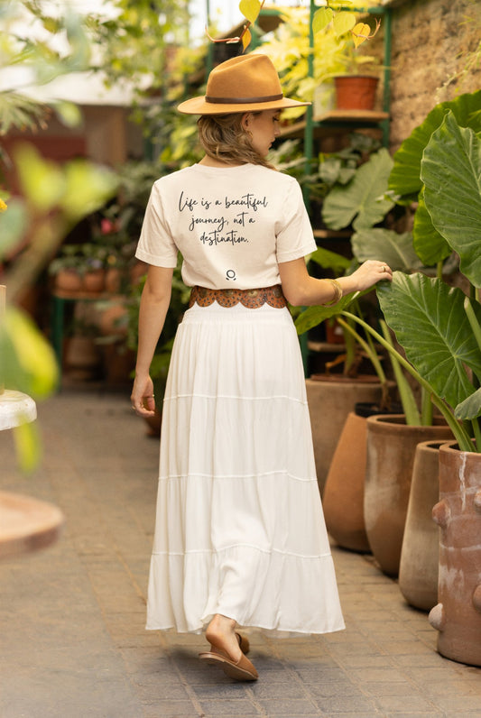 A woman walks down a plant-filled corridor, barefoot in an eco-conscious Bella Vita Voyager Tee, featuring the phrase "Life is a beautiful journey, not a destination." Her tan hat and flowing white skirt complete the peaceful scene as she moves among large potted plants and clay pots. It's truly a Bella Vita Voyageur experience.