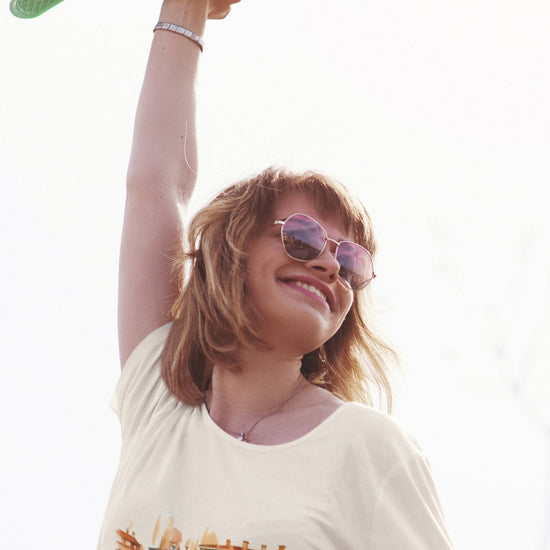 A smiling individual wearing sunglasses raises a green skateboard with one hand, showcasing their white Bella Vita Voyager tee adorned with a colorful urban scene, crafted from organic cotton. The bright and slightly overexposed background adds a sunny and vibrant touch to the moment, ideal for the Bella Vita Voyageur line.
