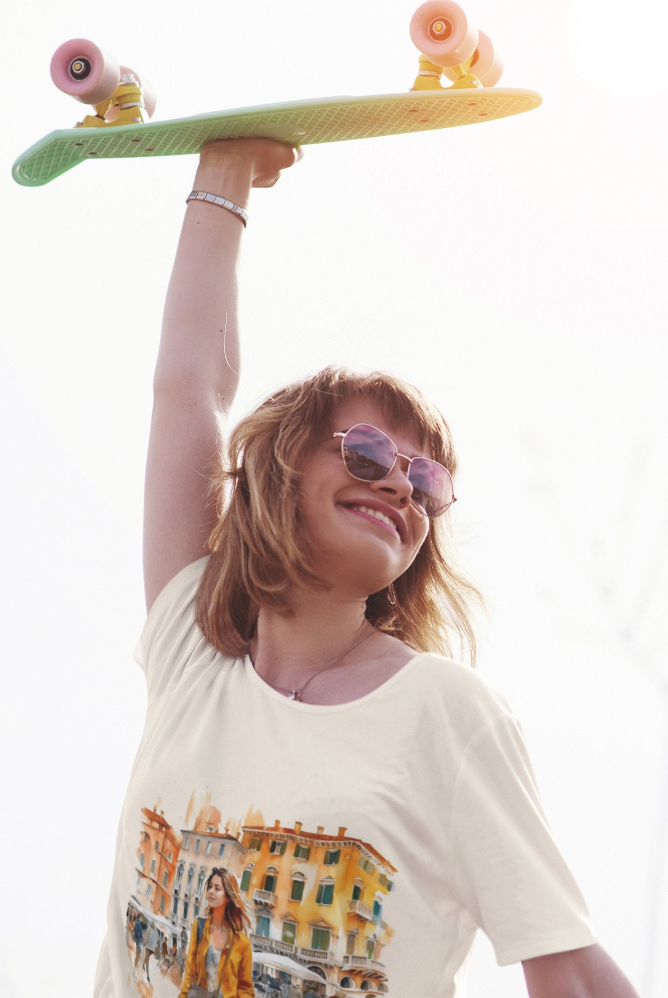 A smiling individual wearing sunglasses raises a green skateboard with one hand, showcasing their white Bella Vita Voyager tee adorned with a colorful urban scene, crafted from organic cotton. The bright and slightly overexposed background adds a sunny and vibrant touch to the moment, ideal for the Bella Vita Voyageur line.