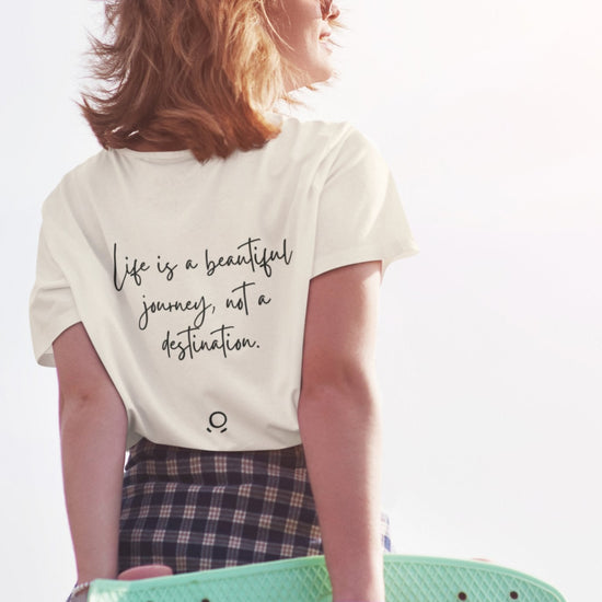A person with short hair holds a turquoise skateboard behind their back. They wear a Bella Vita Voyager Tee in white, which features the quote, "Life is a beautiful journey, not a destination," along with a plaid skirt. The person smiles as they gaze toward the sunlight, creating an uplifting and vibrant scene.