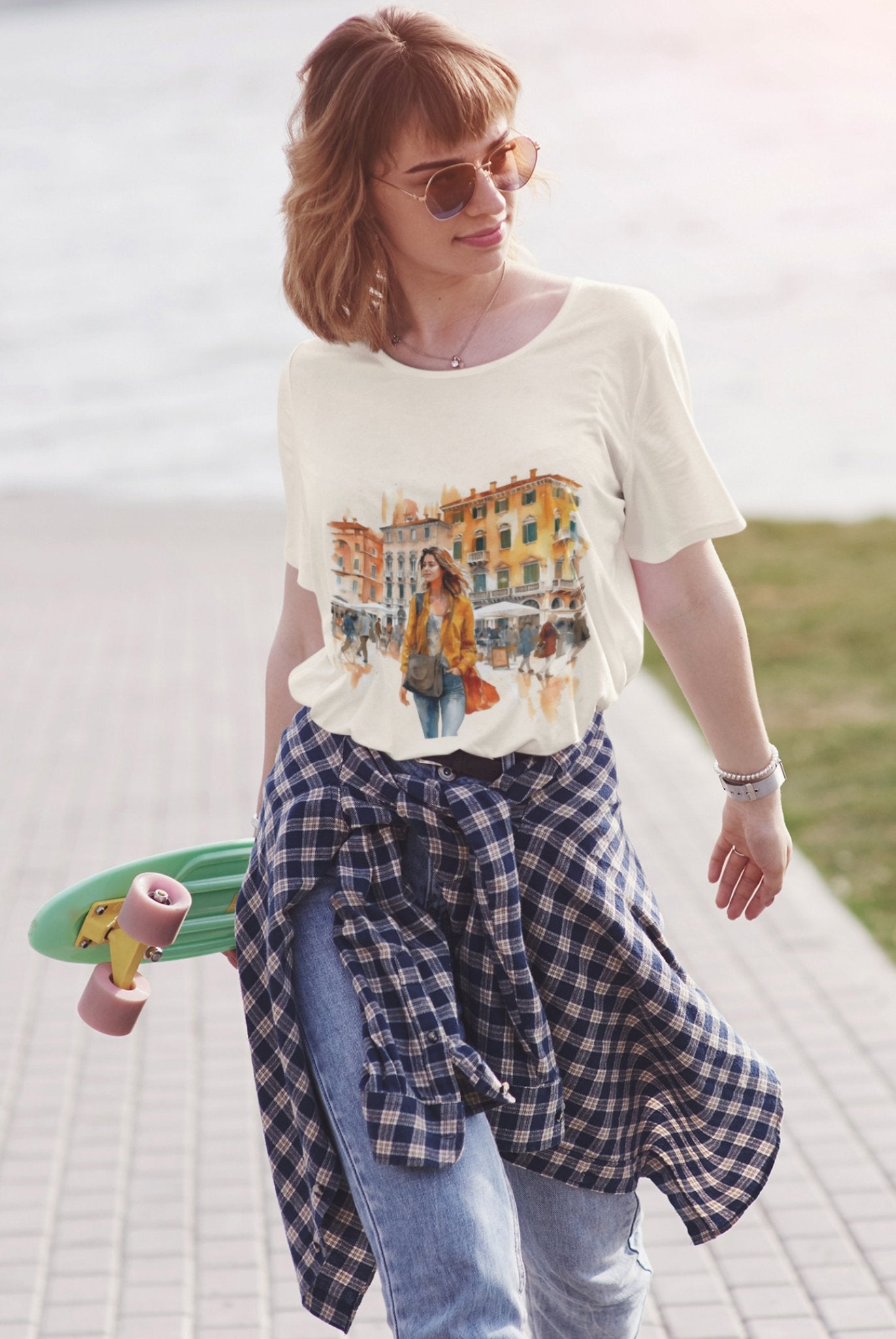 A woman strolls outdoors with a skateboard, wearing sunglasses, a Bella Vita Voyager Tee, jeans, and a plaid shirt tied at her waist. Her shoulder-length hair frames a soft smile. In the background, blurred water and a pathway hint at her adventurous spirit.