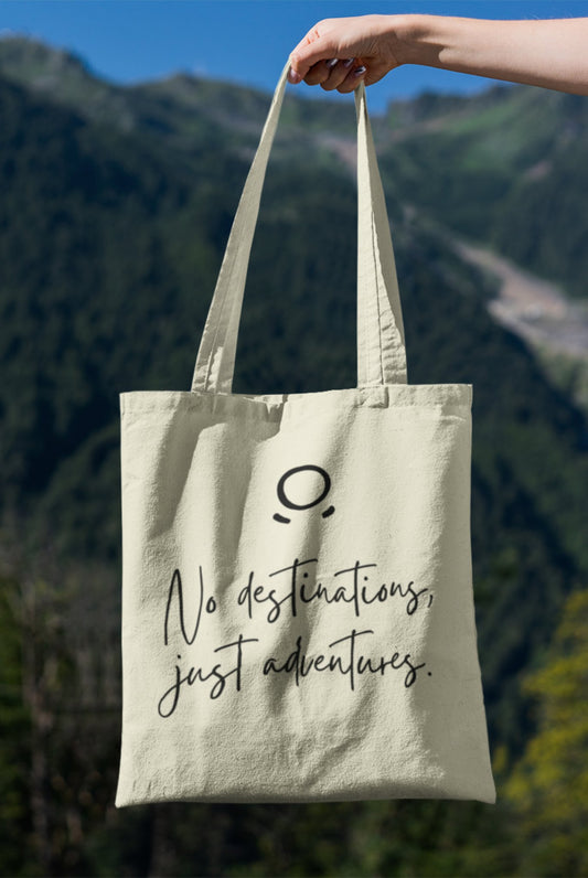 A hand holds the Adventure Awaits Tote Bag against a mountain backdrop. This eco-friendly tote bag showcases an elegant minimalist design with a small circle above a curved line, resembling a face, and is inscribed with the cursive message "No destinations, just adventures." Adventure Awaits!