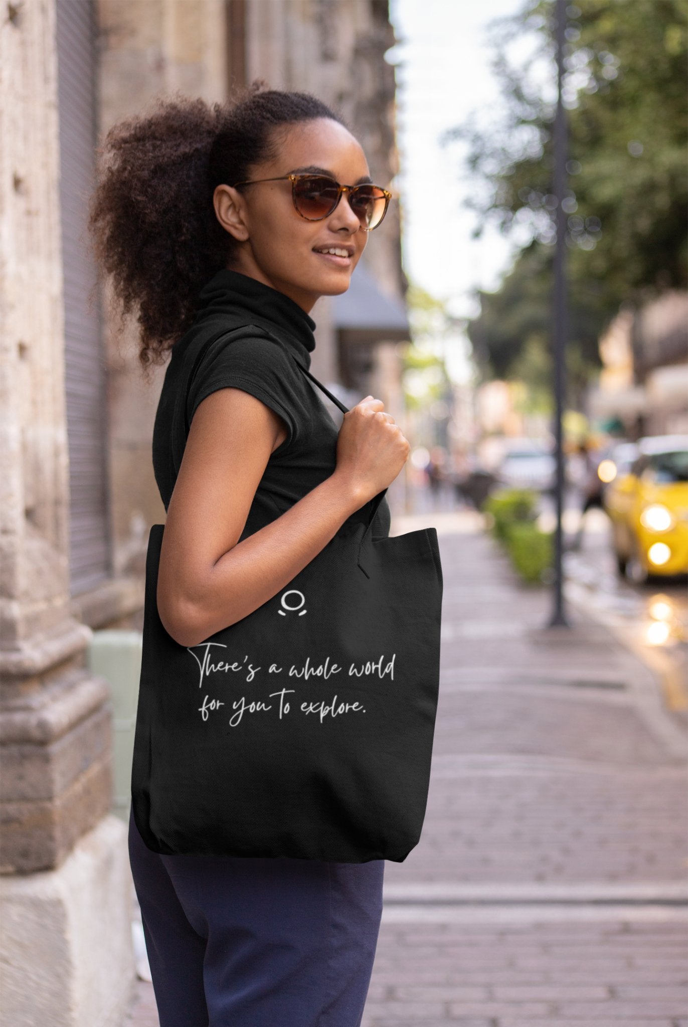 A woman with sunglasses and curly hair stands on a city sidewalk, smiling brightly. Embracing her adventurous spirit, she dons a black sleeveless top and blue pants while carrying an eco-friendly Cosmic Voyager Tote Bag emblazoned with the message, "There's a whole world for you to explore." Behind her, trees line the street where a yellow car is parked.