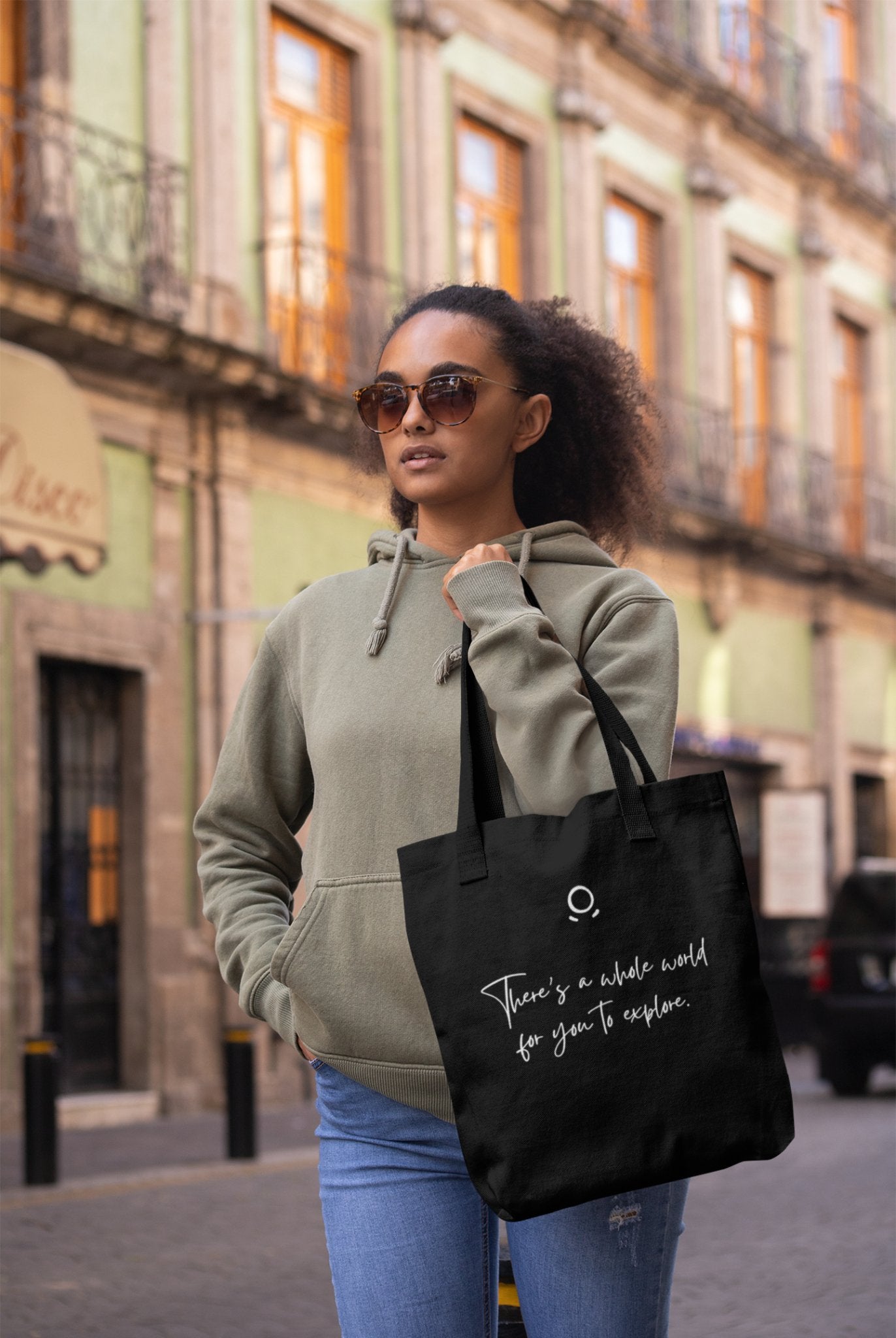 A woman with an adventurous soul, wearing sunglasses and a green hoodie, holds a Cosmic Voyager Tote Bag that reads "There's a whole world for you to explore." She stands on a city street with historic buildings in the background, featuring ornate balconies and large windows.