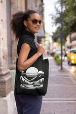 A woman wearing sunglasses and a black turtleneck smiles as she strolls down a city street, carrying the Cosmic Voyager Tote Bag. This eco-friendly tote showcases a white graphic of a mountainous landscape with trees, a river, and a large sun or moon. In the background, there is a blurred yellow car and some greenery.