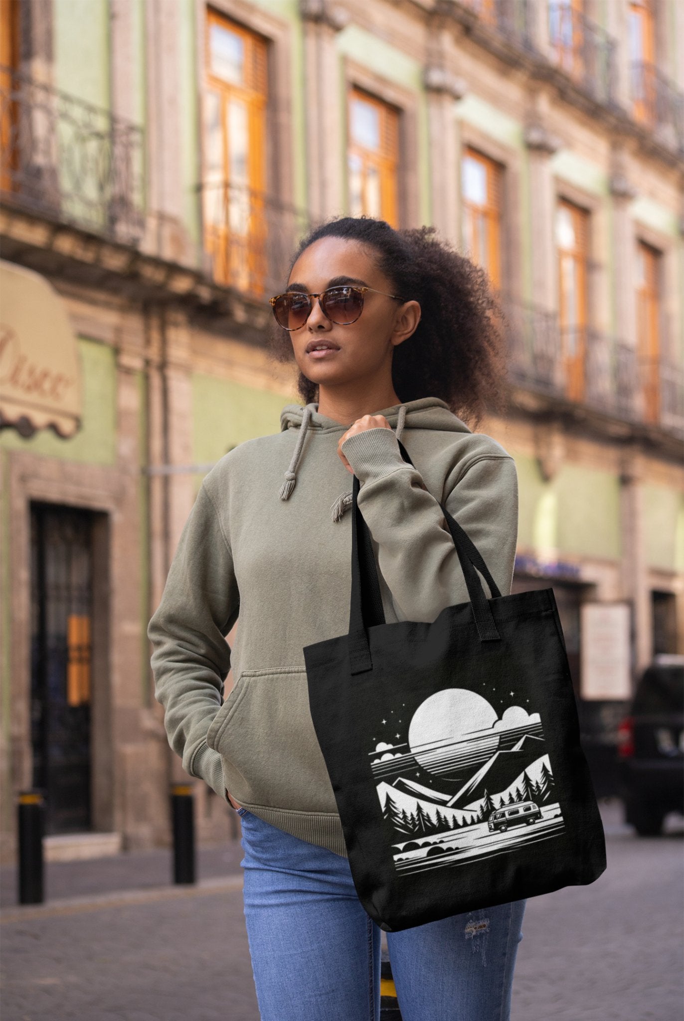 A person wearing sunglasses and a green hoodie strolls down a city street, embodying an adventurous soul. They carry the Cosmic Voyager Tote Bag, adorned with mountains, trees, and a moon. Behind them, a building with green walls and ornate balconies completes the scene.