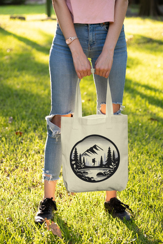 A person stands on grass holding the Adventure Awaits Tote Bag, featuring a mountain and forest silhouette design. They wear a light pink top, ripped blue jeans, black sneakers, and a watch. The sun casts long shadows, suggesting late afternoon—perfect for sustainable exploration.