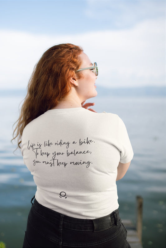 A woman with long red hair stands by a tranquil lake, facing away from the camera. She wears sunglasses and a Pedal through Life Tee, crafted from organic cotton for an eco-friendly touch. The shirt displays the text, "Life is like riding a bike. To keep your balance, you must keep moving." Above her is a clear sky, and in front of her stretches a wooden dock into the water.