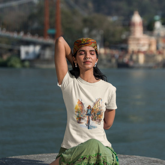 A person sits cross-legged on a stone ledge by a river, performing a yoga pose with one arm raised. They wear a colorful headband, the eco-friendly "Pedal through Life Tees," and a green skirt. In the background, there's a suspension bridge and blurred buildings amid a hilly landscape.